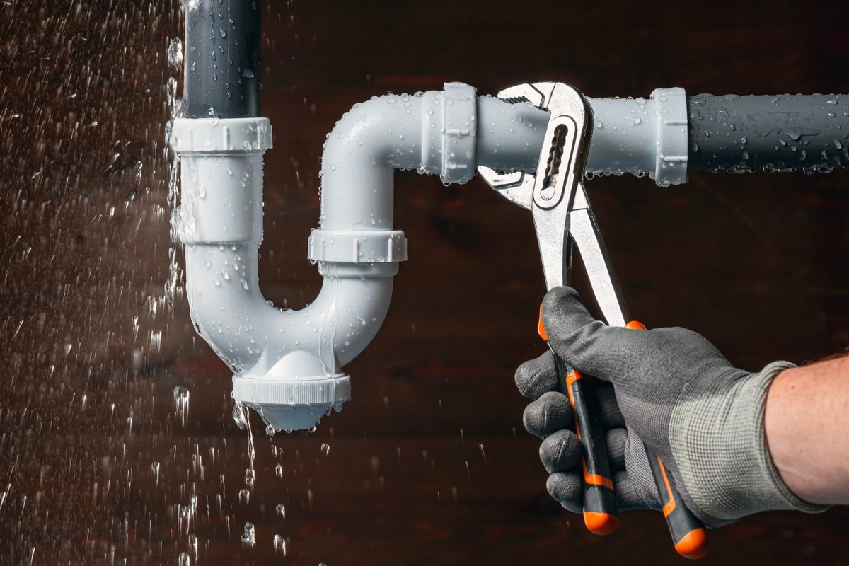 Gloved hand tightening a leaking sink drain trap with pliers as water sprays out